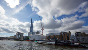 This urban photograph captures an Uber Boat by Thames Clippers traveling along the River Thames in London on a summer morning. The image features the distinctive architecture of the city’s riverside buildings, including the prominent landmark The Shard, which rises sharply into the sky behind the boat. Overhead, a partially clouded sky is illuminated by the soft summer morning light, while a construction crane can be seen working near the modern buildings along the riverbank. The scene exemplifies London’s dynamic mix of contemporary and historic architecture along the Thames, set against a busy cityscape with the Uber Boat serving as the main subject.